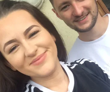 Two people smiling; the person on the left has long dark hair and eyebrow styling, and the person on the right has facial hair.