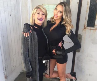 Two women smiling and posing together, one wearing a patterned coat and the other in a black outfit, against a rustic backdrop.