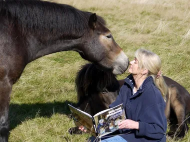 A woman sits on grass with a book titled "Wild Pony Whispering," gently interacting with a pony nuzzling her face.