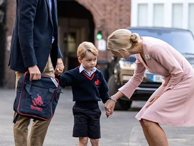 Child in school uniform holds hands with an adult outside a building, with another adult in pink bending down to greet him.