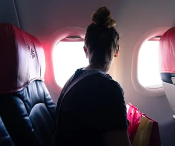 Passenger looking out airplane window, seated next to pink and black seats, holding shopping bags.