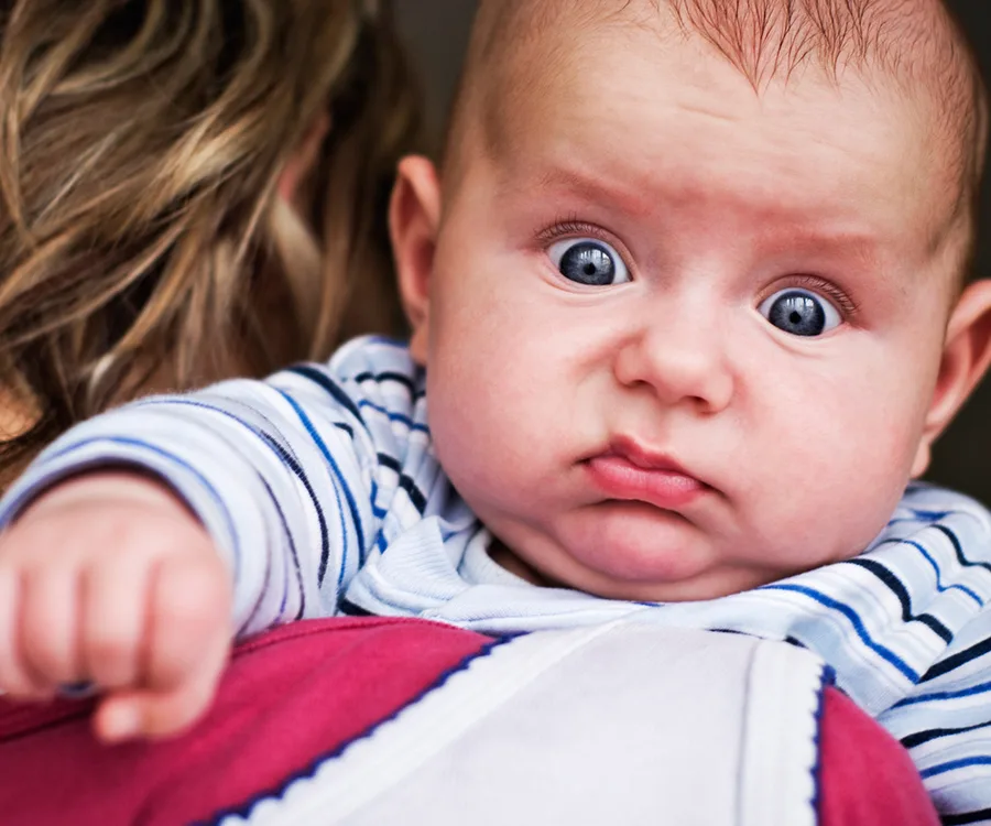 Close-up of a baby with a curious expression, wearing a striped outfit, being held by a person with blonde hair.