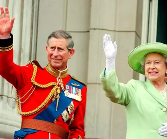 Prince in red uniform and medals, with a woman in green suit and hat, both waving on a balcony.