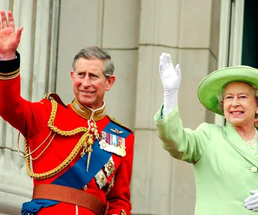 Prince in red uniform and medals, with a woman in green suit and hat, both waving on a balcony.