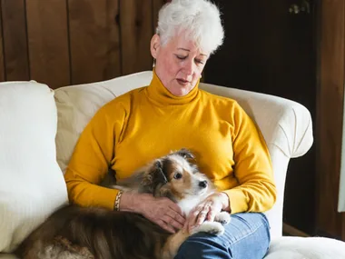 Woman in a yellow sweater sitting on a couch, gently holding a small dog in her lap.