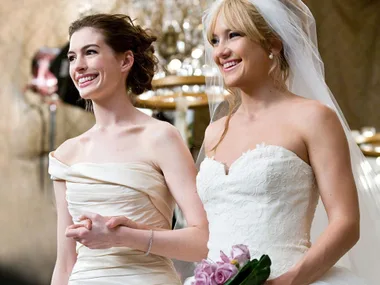 Two brides smiling, holding hands, in bridal dresses with bouquet. Scene from the movie "Bride Wars."