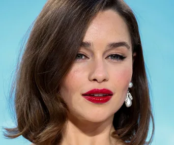 A person with medium brown hair, red lipstick, and pearl earrings against a light blue background.