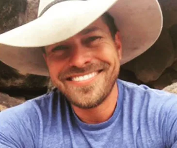 Man in a blue shirt and wide-brimmed hat smiling outdoors with rocks in the background.