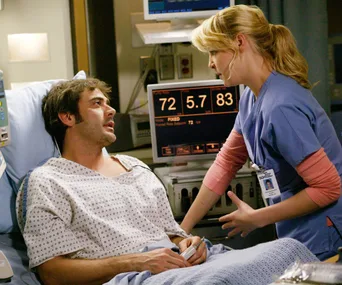 A doctor in scrubs tends to a patient lying in a hospital bed. Medical monitors are visible in the background.