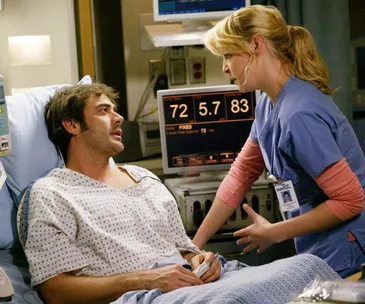 A doctor in scrubs tends to a patient lying in a hospital bed. Medical monitors are visible in the background.