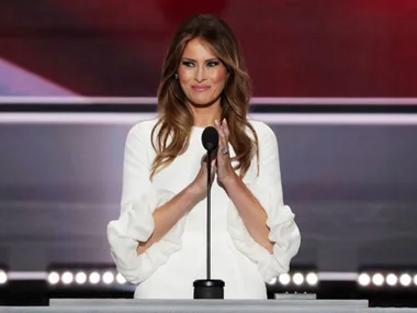 A woman in a white dress stands at a podium, clapping with a smile against a blurred background.