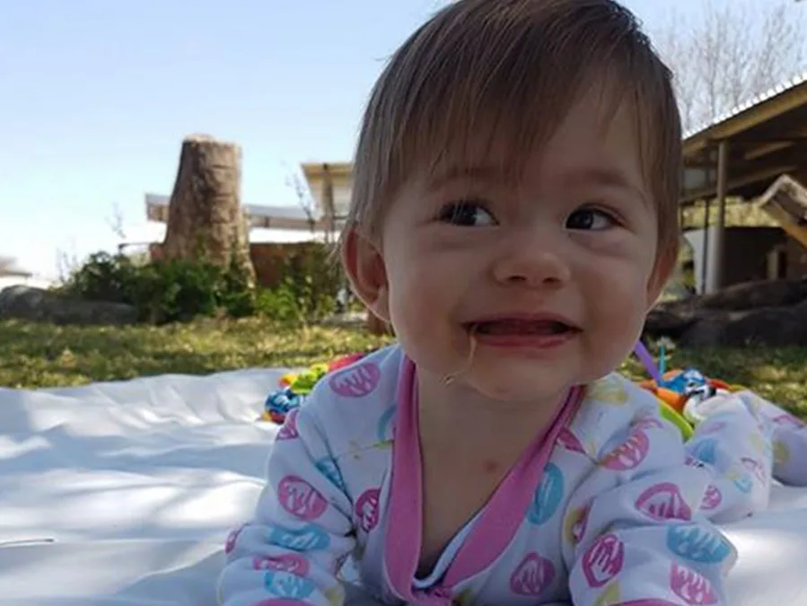 A smiling baby lying on a blanket outside, wearing a colorful patterned onesie, with grass and trees in the background.