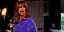 A woman with long hair in a blue sweater, angrily yelling in a dimly lit room.
