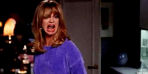 A woman with long hair in a blue sweater, angrily yelling in a dimly lit room.