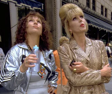 Two women look up; one in a silver jacket holds a bottle, the other in a snakeskin coat holds sunglasses on a city street.