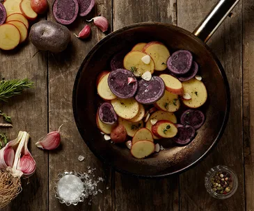 Sliced purple and red potatoes with garlic and herbs in a pan on a wooden surface, surrounded by ingredients.