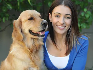 A woman smiling alongside a Golden Retriever, with greenery in the background.