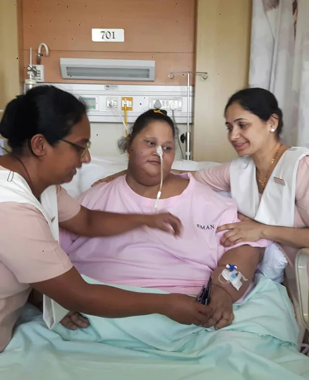 A patient in a hospital bed, wearing a pink gown, is assisted by two medical staff members.