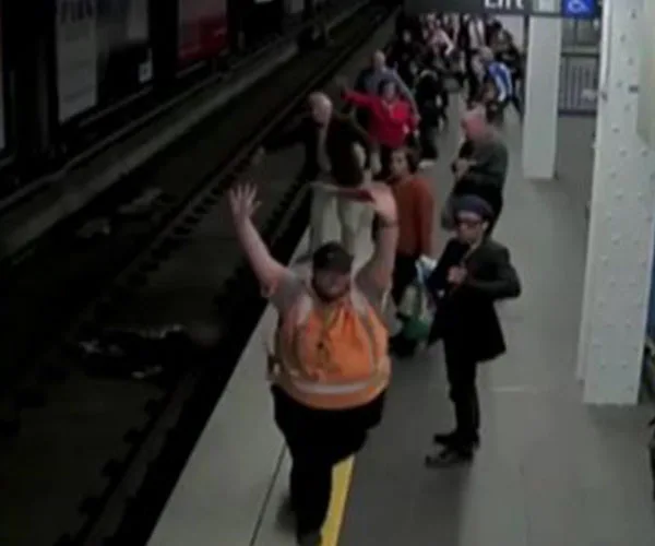 A crowded train station platform with people near the edge, one person in an orange vest raising their arms.