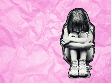 A young girl sits with her head on her knees, looking sad, on a crumpled pink paper background.