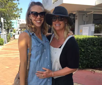 Two women smiling outdoors, one is pregnant. Both wearing sunglasses and embracing on a sunny day.