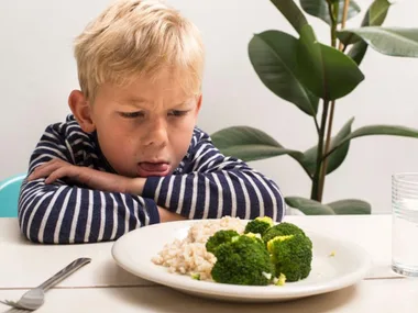 Child in striped shirt grimacing at a plate of broccoli and rice, sitting at a table with a plant in the background.