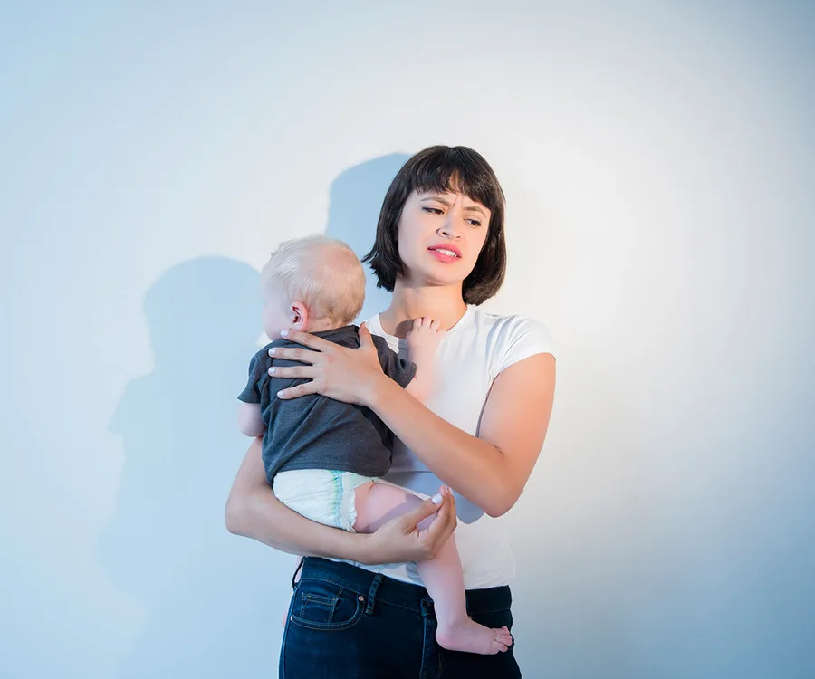 A woman holds a baby with a concerned expression against a plain background.