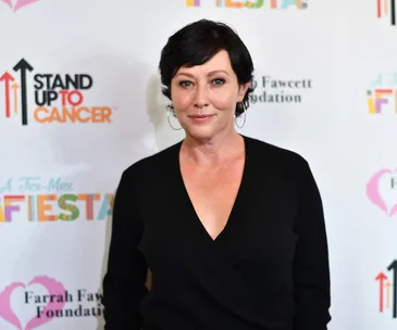 Shannen Doherty at a Stand Up To Cancer event, wearing a black top, with event branding in the background.