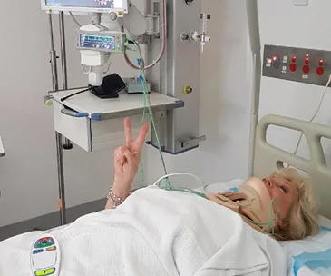 Patient lying in a hospital bed giving a peace sign, with medical equipment in the background.