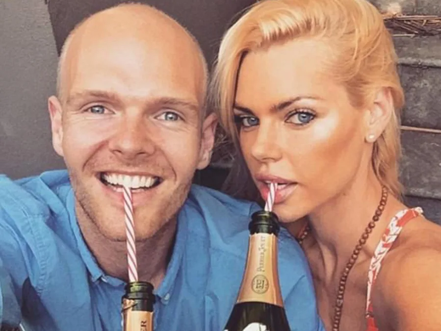 A woman and man sip from champagne bottles with striped straws, smiling at the camera.