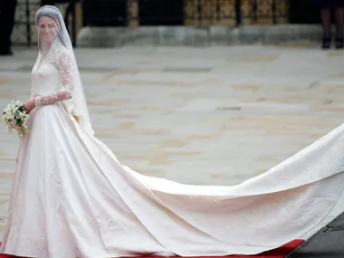 Bride in lace wedding dress and long veil smiling, holding bouquet on red carpet outdoors.