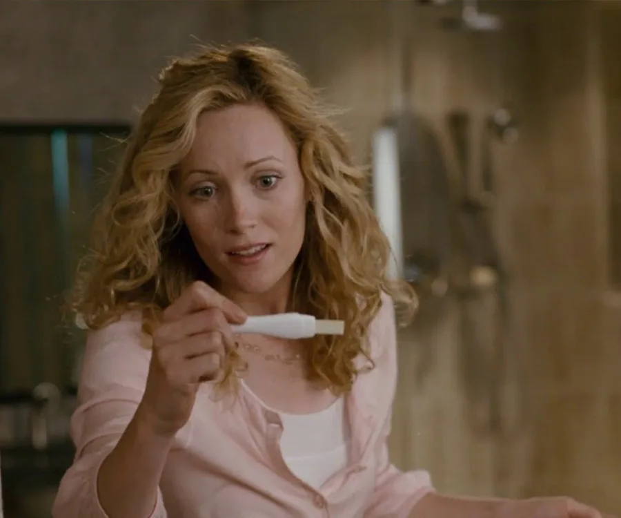 A woman with curly hair examines a pregnancy test with a neutral expression in a bathroom setting.