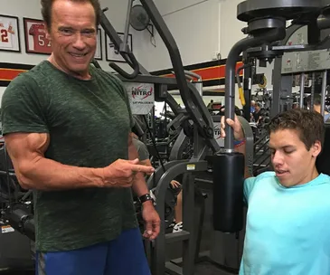Arnold Schwarzenegger points at young man using gym equipment, smiling, with exercise machines in background.