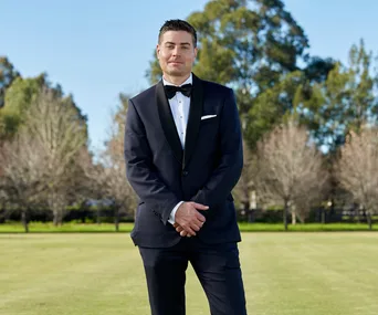 Man in a suit stands on grass with trees in the background under a clear sky.