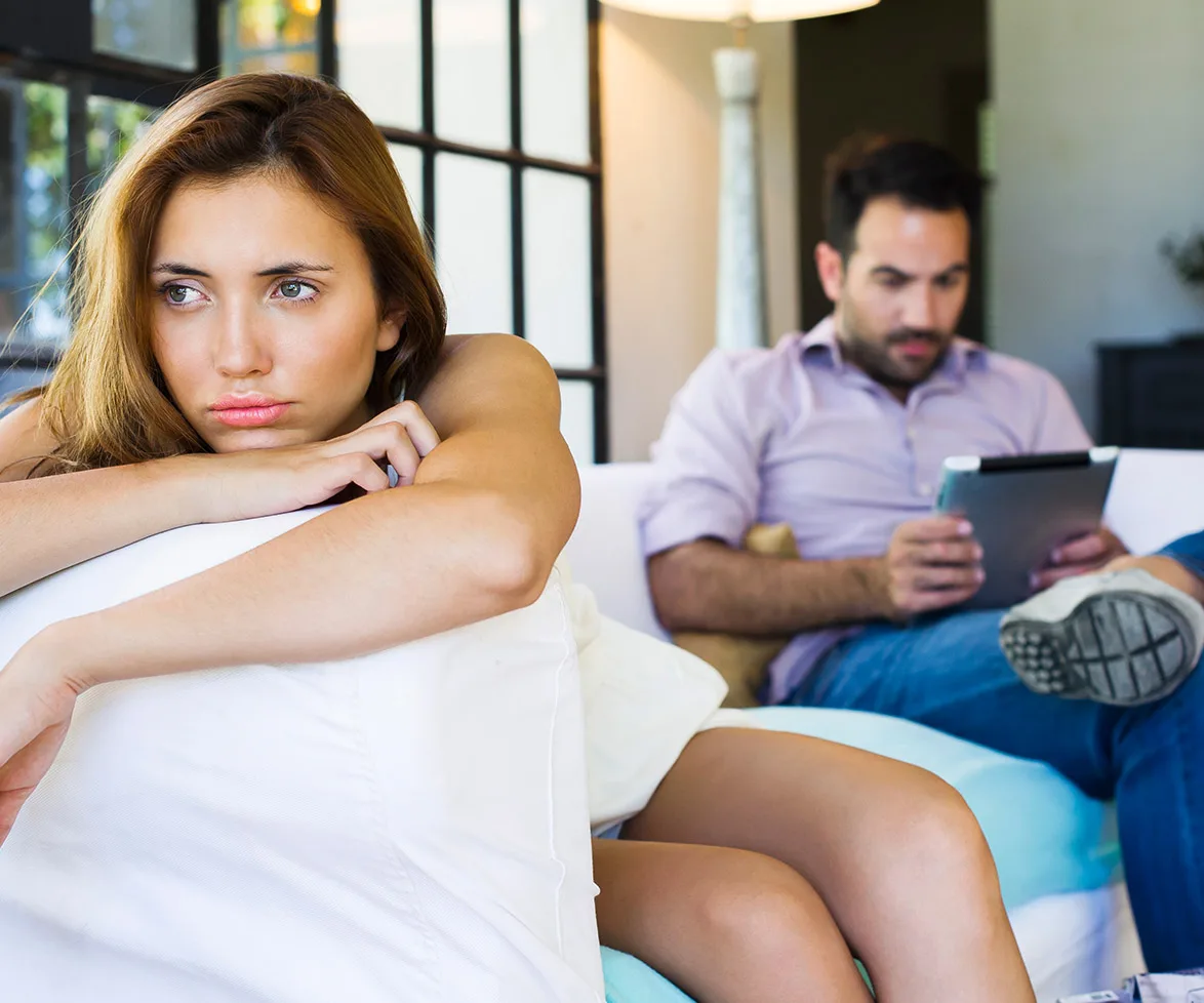 Woman looks upset on sofa while man in background is focused on a tablet.