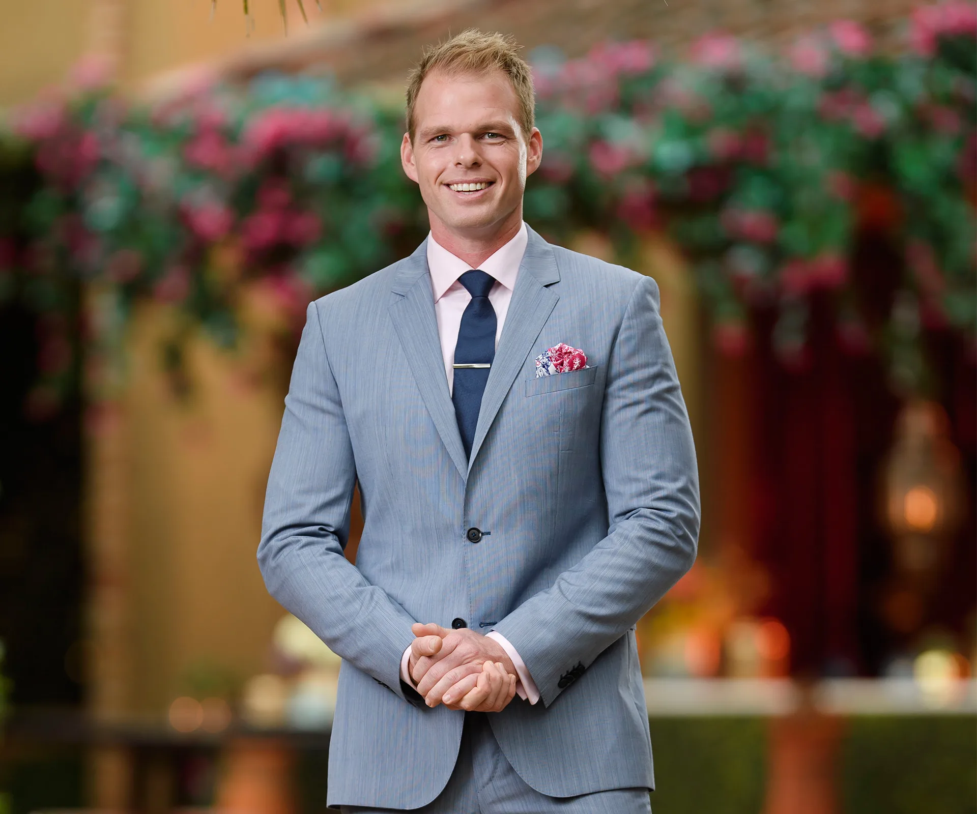 Man in a blue suit with a pink shirt and navy tie, smiling in a garden setting with colorful flowers in the background.