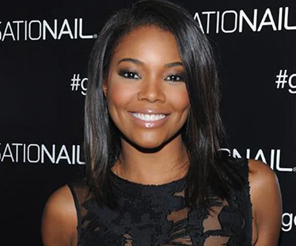 Gabrielle Union smiling at an event, wearing a black lace dress with a branded backdrop behind her.