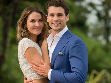 A couple smiling outdoors, the man in a blue suit and the woman in a white top, embracing each other.