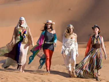 Four women in colorful outfits walking through a desert landscape, showcasing fashion and camaraderie.