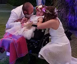 A couple kisses a child in a wheelchair wearing a dress and flower crown during a touching outdoor wedding ceremony.