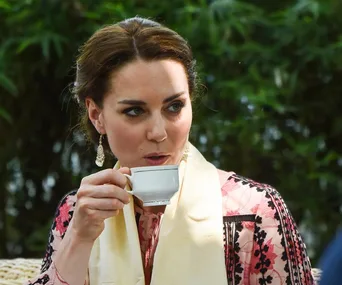 A person sipping tea outdoors, wearing a patterned outfit with earrings and a scarf, surrounded by greenery.