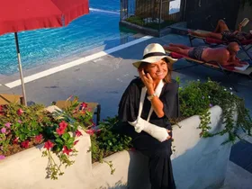 Woman with arm in sling smiling by poolside, wearing a sunhat, beside flowers and under an umbrella.
