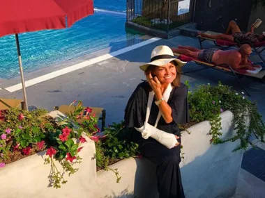 Woman with arm in sling smiling by poolside, wearing a sunhat, beside flowers and under an umbrella.