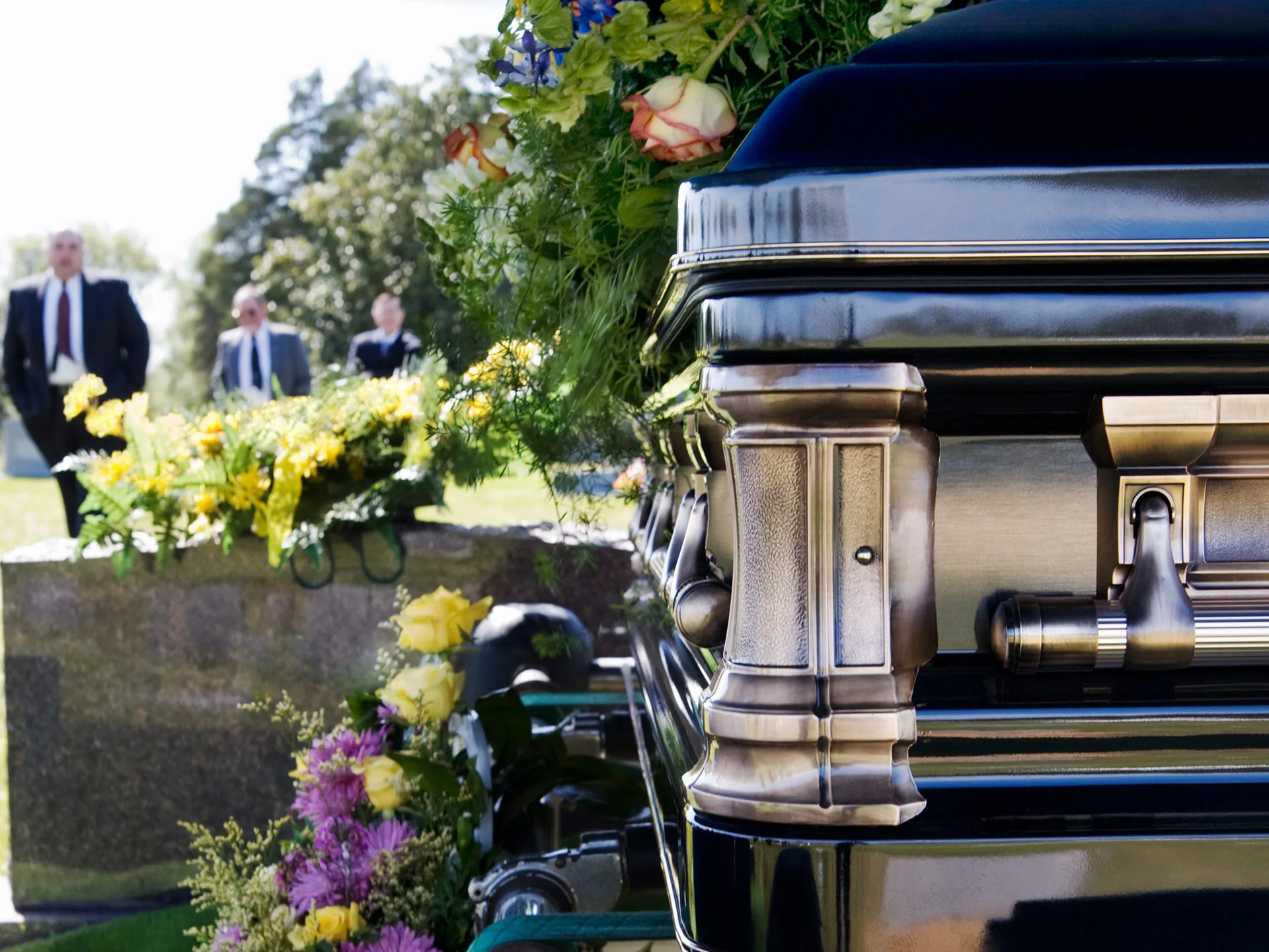 A polished casket with floral arrangements, and people in suits standing in the background at a cemetery.