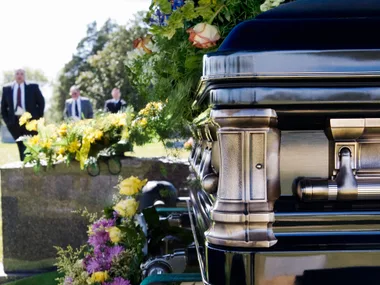A polished casket with floral arrangements, and people in suits standing in the background at a cemetery.
