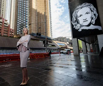 Woman in a cityscape stands confidently beside a large black and white portrait on a building.