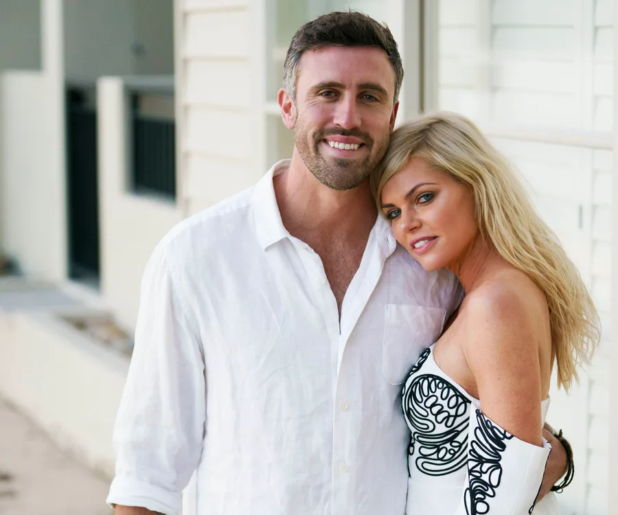 A couple posing closely; the man in a white shirt smiles, while the woman leans on him, wearing a dress with black details.