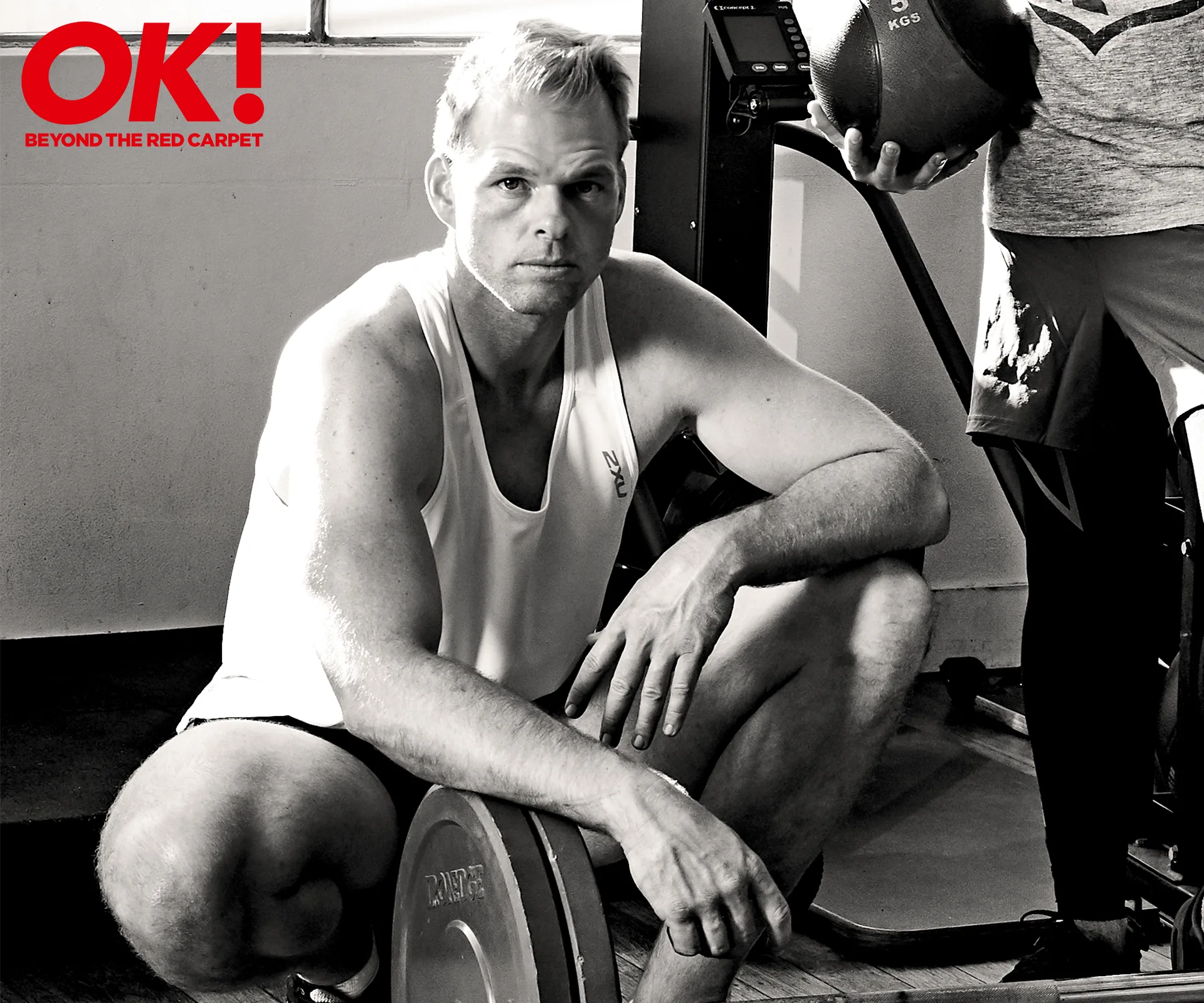 Person in a gym, crouching by weights, wearing a white tank top.