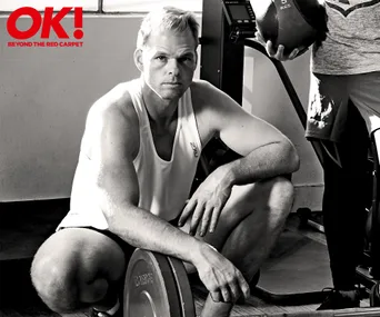 Person in a gym, crouching by weights, wearing a white tank top.