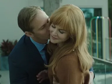 A man in a suit hugs and kisses a smiling woman with red hair, outdoors by a playground.